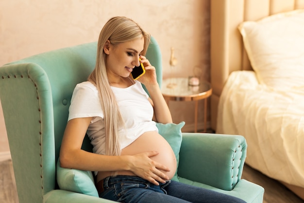 Mujer adulta joven embarazada sentada en una silla azul y hablando por teléfono.