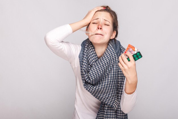 La mujer adulta joven enferma tiene temperatura y tiene muchas pastillas. Foto de estudio, aislado sobre fondo gris