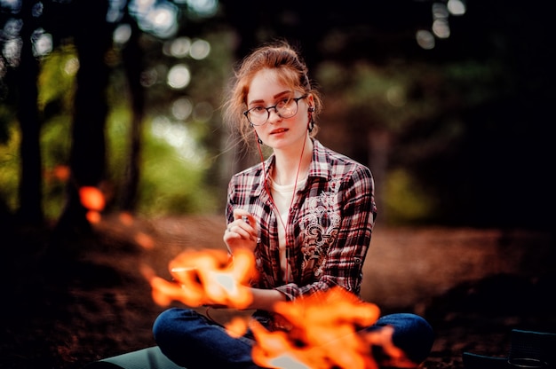 Mujer adulta joven de pelo rojo con gafas y auriculares disfrutando de escuchar música en el bosque cerca del fuego