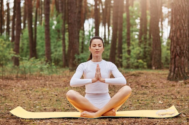 Mujer adulta joven sentada en el suelo en karemat en posición de loto, manteniendo las palmas juntas, posando con los ojos cerrados, relajándose y meditando al aire libre en el bosque, yoga en la naturaleza.