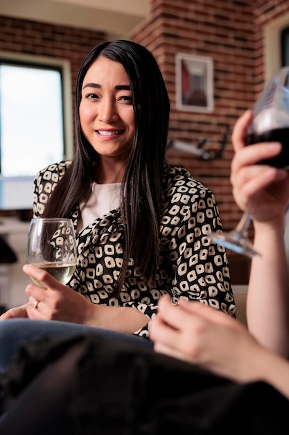 Mujer adulta joven sonriente de corazón bebiendo vino en la fiesta de cumpleaños mientras mira la cámara. Hermosa chica disfrutando pasar el rato con amigos en casa mientras celebra un momento importante de la vida.