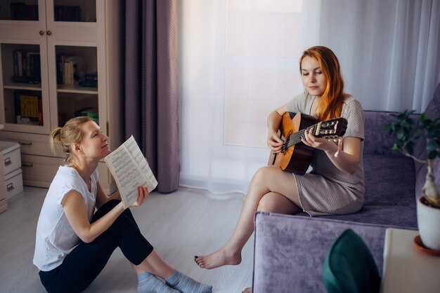 Mujer adulta joven sostiene el cuaderno de música y mira a su amiga tocando la guitarra. Dos chicas lindas tocan música en casa. Amistad, amor, ocio familiar, aficiones.