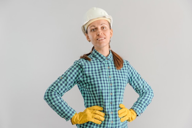 Una mujer adulta joven trabajador de la construcción en un casco de construcción blanco está parado con sus manos en guantes amarillos en sus caderas sobre un fondo gris