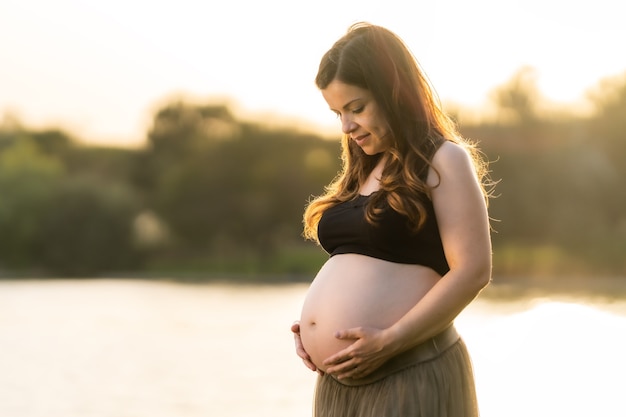 Mujer adulta latina embarazada mirando y tocando su abdomen con una falda cómoda mientras está de pie junto a un lago en un parque mientras el sol brilla sobre ella