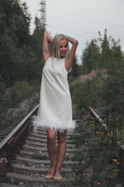 Mujer adulta en la naturaleza caminando en el bosque de verano en el ferrocarril del atardecer. Señora soñadora en vestido caminar por la noche en el ferrocarril. Emoción positiva femenina sobre raíles al amanecer