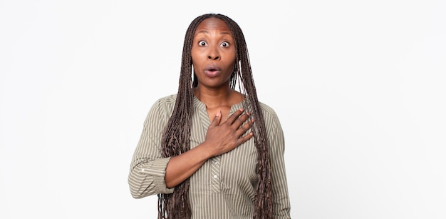 Mujer adulta negra afro que se siente consternada y sorprendida, sonriendo, tomando la mano al corazón, feliz de ser la indicada o mostrando gratitud