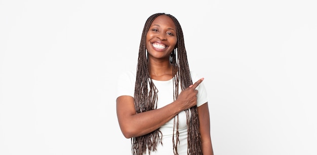 Mujer adulta negra afro sonriendo alegremente, sintiéndose feliz y apuntando hacia un lado y hacia arriba, mostrando el objeto en el espacio de la copia