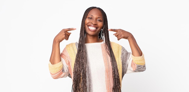 Mujer adulta negra afro sonriendo con confianza apuntando a su propia sonrisa amplia, actitud positiva, relajada y satisfecha