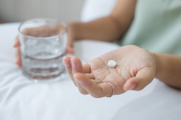 Mujer adulta con píldora y vaso de agua, tomando medicamentos en la cama por la mañana en su casa. Concepto de migraña, analgésico, dolor de cabeza, gripe, enfermedad, enfermedad y atención médica