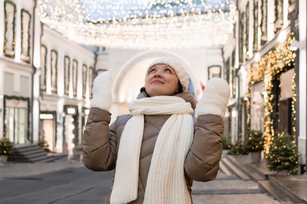 Una mujer adulta en ropa de invierno en la ciudad mira la iluminación navideña y admira lo que ve.