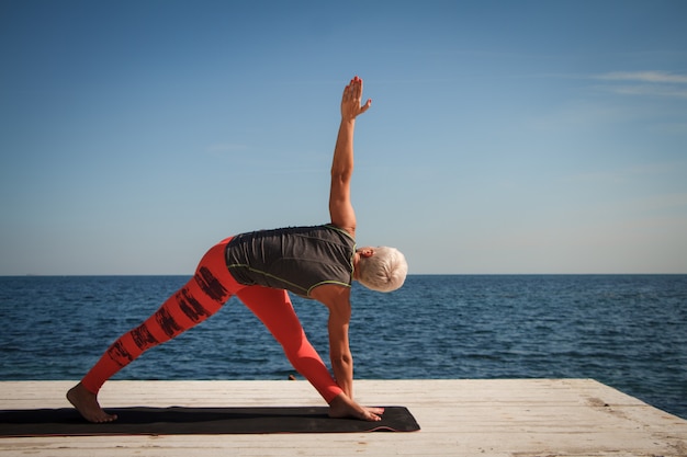 Mujer adulta rubia con corte de pelo corto practica yoga en el muelle contra el fondo del mar y el cielo azul