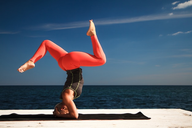 Mujer adulta rubia con corte de pelo corto practica yoga en el muelle contra el fondo del mar y el cielo azul