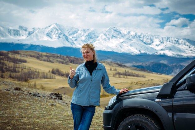 La mujer adulta sonriente alegre se coloca en una colina cerca del coche contra la cordillera nevada. Viajero de la mujer al aire libre en un día soleado que muestra el pulgar hacia arriba.