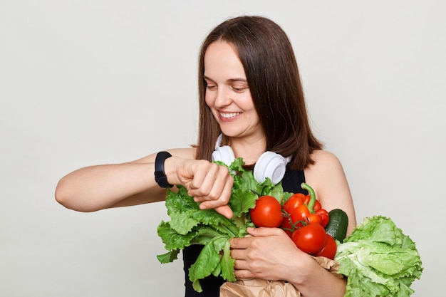 Mujer adulta sonriente con cabello castaño aislada sobre fondo blanco sosteniendo verduras mirando su rastreador de fitness contando calorías quemadas