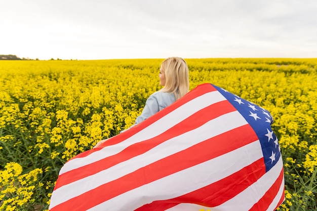 Mujer adulta sosteniendo la bandera americana con poste, estrellas y rayas en un campo de colza amarillo. Bandera de Estados Unidos ondeando en el viento.