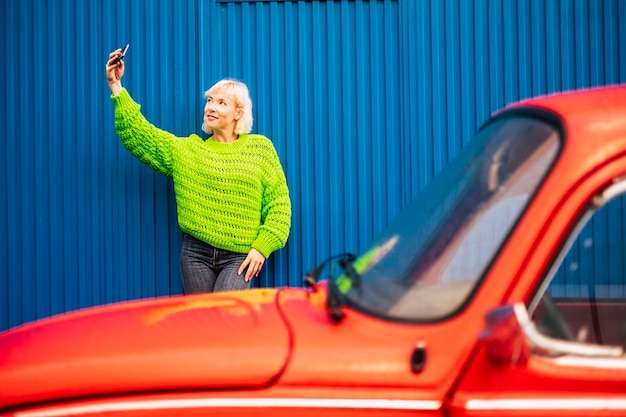 Foto mujer adulta tomando una selfie contra el hierro corrugado con un coche rojo en primer plano