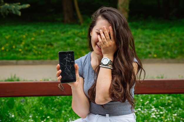 Mujer adulta triste joven que muestra el teléfono agrietado