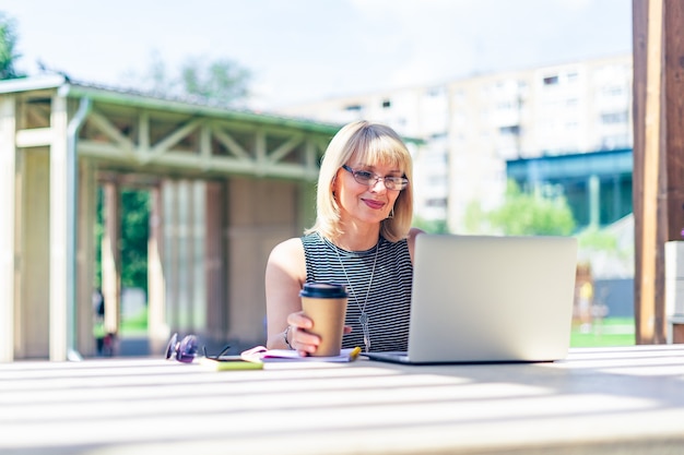 Mujer adulta en vidrios con videollamada con laptop afuera en el parque. Feliz y sonriente senior trabajando y tomando café.
