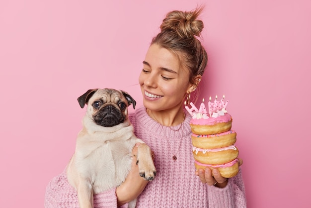 Una mujer afectuosa y alegre mira con amor al perro pug celebra su cumpleaños sostiene dulces donuts con velas encendidas cuida de su mascota favorita usa un suéter de punto aislado sobre un fondo rosa