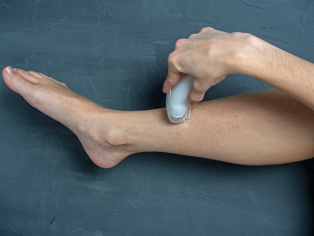 Una mujer se afeita la pierna con una depiladora eléctrica sobre un fondo de textura gris. El concepto de belleza y cuidado personal.