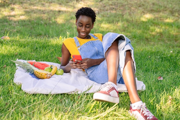 Mujer africana acostada en un parque al aire libre charlando y enviando mensajes de texto en su teléfono celular mientras hace un picnic