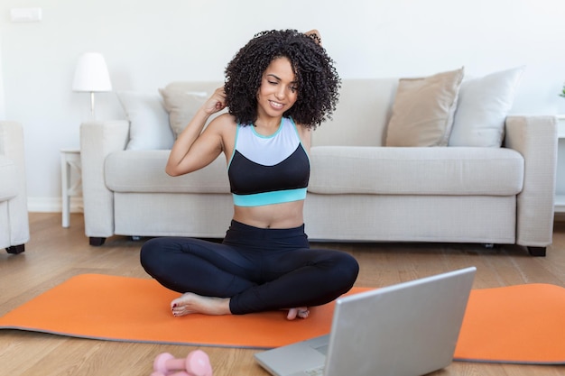 Una mujer africana adulta hace ejercicios de yoga y entrenamiento de fuerza en una alfombra en su sala de estar Sigue un video de un curso de ejercicios en línea en su computadora portátil