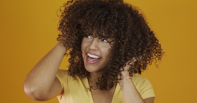 Mujer africana americana riendo feliz con su pelo rizado sobre fondo amarillo. Riendo a mujer rizada en suéter tocando su cabello y mirando a la cámara.