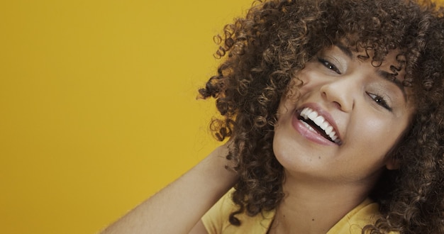 Mujer africana americana riendo feliz con su pelo rizado sobre fondo amarillo. Riendo a mujer rizada en suéter tocando su cabello y mirando a la cámara.