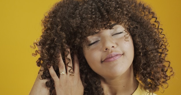 Mujer africana americana riendo feliz con su pelo rizado sobre fondo amarillo. Riendo a mujer rizada en suéter tocando su cabello y mirando a la cámara.