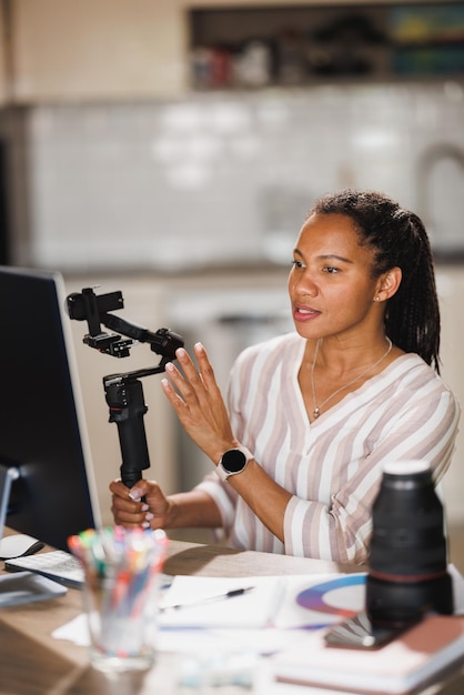 Mujer africana blogueando sobre productos fotográficos y mostrando cardán para cámara mientras se reúne en línea en su computadora en casa.