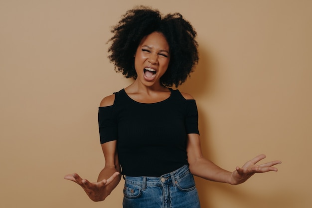 Mujer africana en camiseta negra posando gritando, haciendo las palmas abiertas, sintiéndose enojada, con la boca abierta gritando en voz alta mirando a cámara, aislado sobre fondo beige de estudio. Concepto de emociones negativas