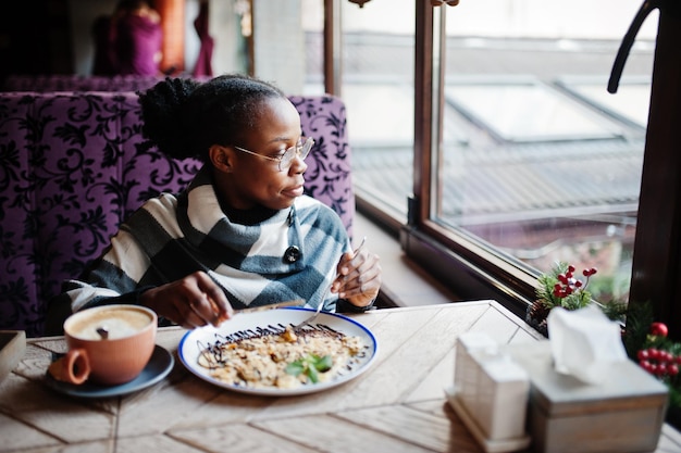 Mujer africana con capa a cuadros y anteojos sentada en el café y comiendo postre