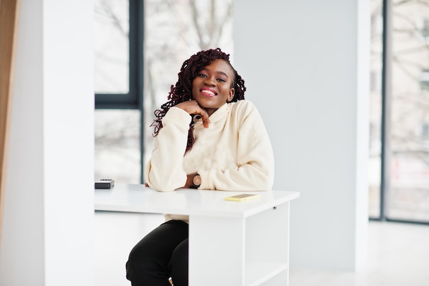 Foto mujer africana con capucha sentada junto a una mesa en un apartamento interior cerca de grandes ventanales.