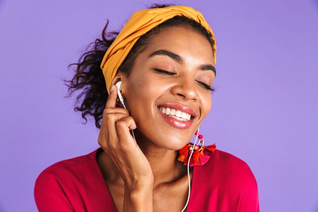 Mujer africana complacida en vestido escuchando música con auriculares con los ojos cerrados sobre la pared púrpura