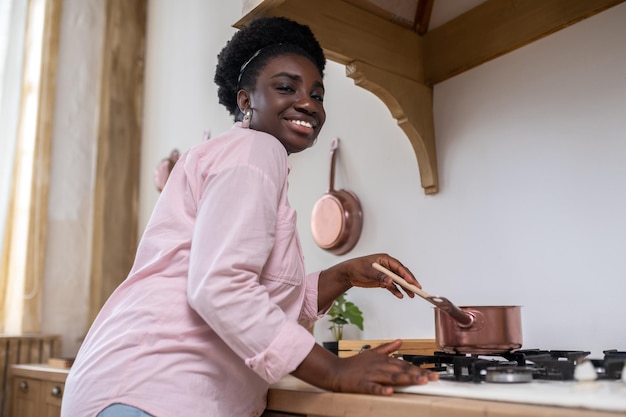 Mujer africana contenta con ropa rosa cocinando en la cocina