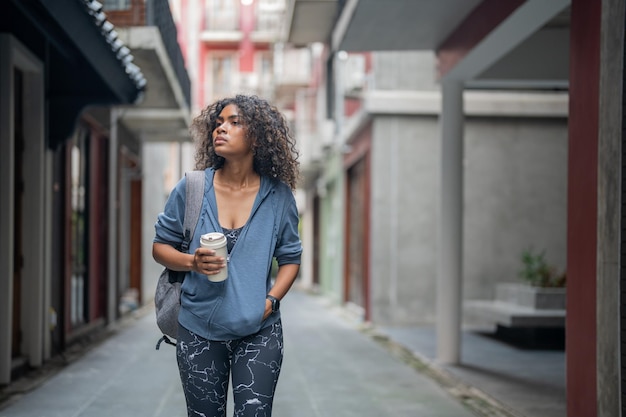 Mujer africana deportiva en forma saludable y firme y delgada bebe agua de una taza en la mano chica fitness con ropa deportiva y chaqueta caminando en la ciudad y el pueblo Fitness y motivación deportiva