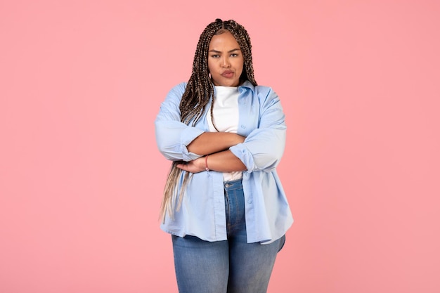 Mujer africana descontenta con sobrepeso posando cruzando las manos sobre fondo rosa