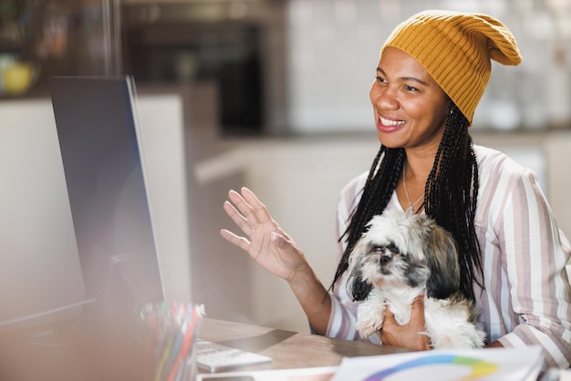 Mujer africana divirtiéndose con su perro mascota y reuniéndose en línea mientras trabaja en la computadora en la oficina de su casa.