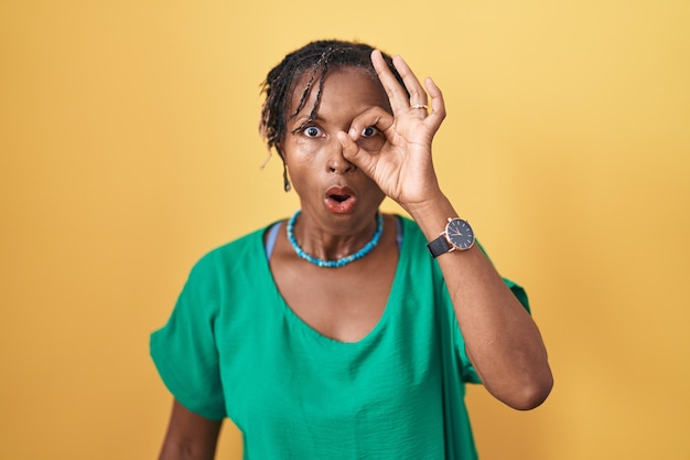 Mujer africana con dreadlocks de pie sobre fondo amarillo haciendo un buen gesto sorprendida con la cara sorprendida, los ojos mirando a través de los dedos. expresión incrédula.