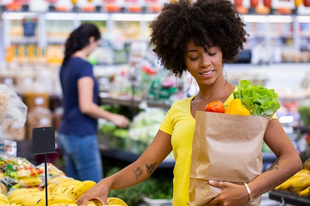 Mujer africana feliz positiva sana sosteniendo una bolsa de papel llena de frutas y verduras.