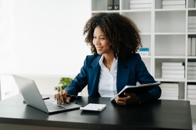 Una mujer africana independiente está trabajando en su trabajo en una tableta y una computadora portátil haciendo análisis de contabilidad, informes de inversión inmobiliaria, datos financieros en la oficina.