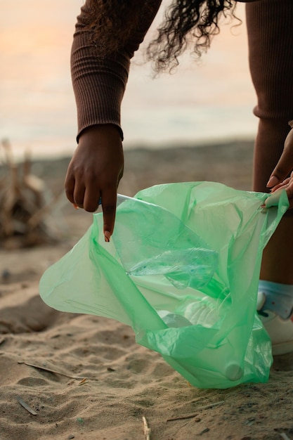 Mujer africana irreconocible inclinándose para recoger basura de la arena en la playa en una bolsa de plástico verde Contaminación ecológica