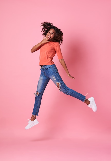 Mujer africana joven alegre en camisa naranja saltando y celebrando sobre fondo rosa.