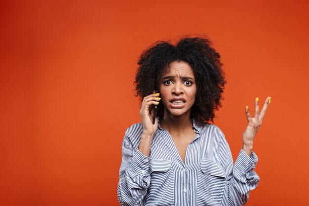 Mujer africana joven enojada hablando por teléfono móvil aislado sobre pared roja