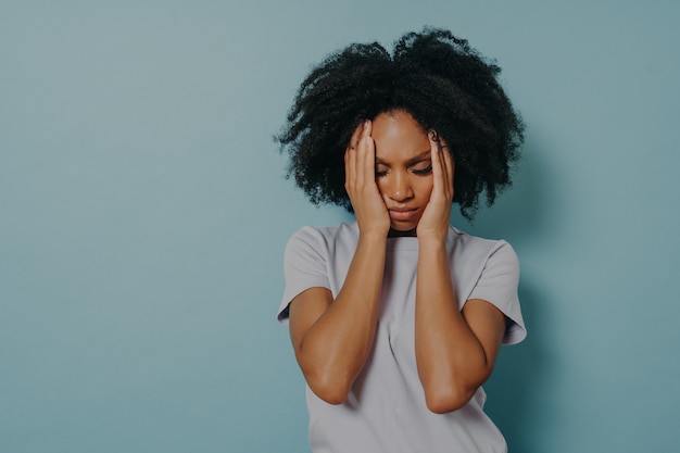 Mujer africana joven estresada cansada en camiseta sosteniendo la cabeza entre las manos y manteniendo los ojos cerrados, suspirando de cansancio o sufriendo de dolor de cabeza mientras posa aislado sobre fondo azul