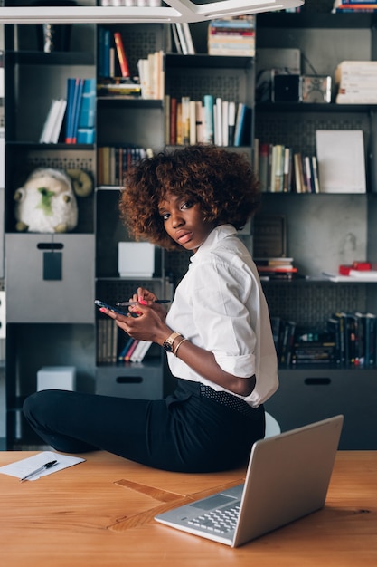 Mujer africana joven que presenta en oficina moderna con smartphone y que mira la cámara