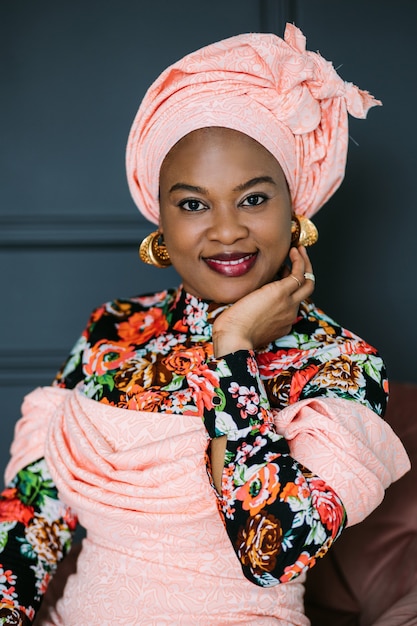 Mujer africana con maquillaje brillante y joyas con estilo, vistiendo un colorido vestido rosa y un pañuelo étnico, posando en el estudio