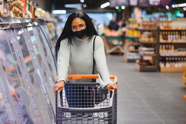 Mujer africana con máscara médica desechable comprando en un supermercado durante el brote de pandemia de coronavirus Tiempo epidémico
