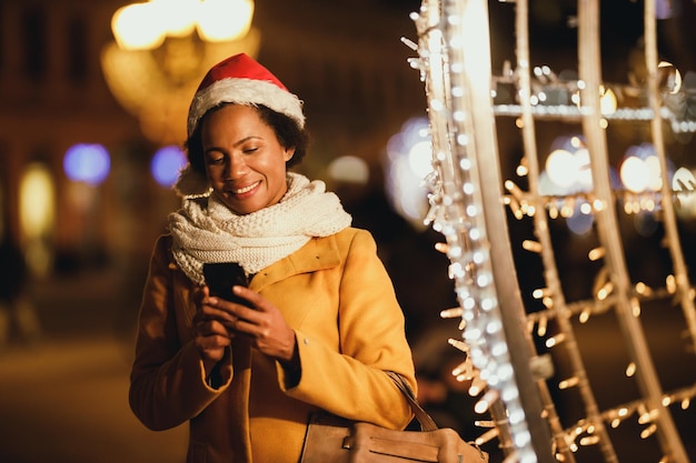 Mujer africana de mediana edad escribiendo en su smartphone en el mercado navideño en la noche festiva de la ciudad.