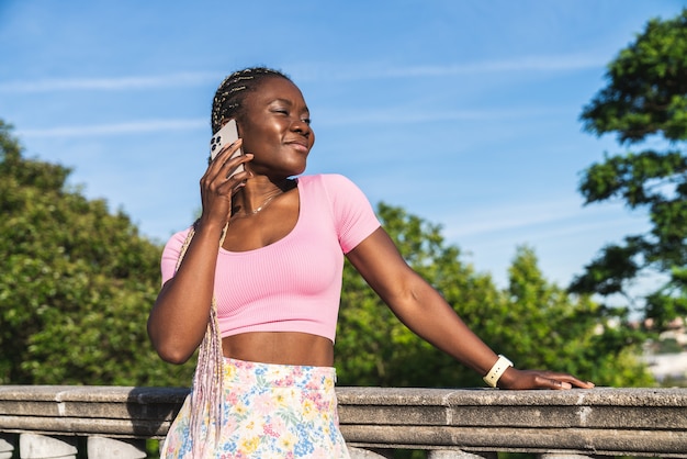 Mujer africana negra hablando por teléfono móvil muy feliz en un parque público en un día muy soleado y cielo gris. Mujer negra en ropa de estilo de vida causal